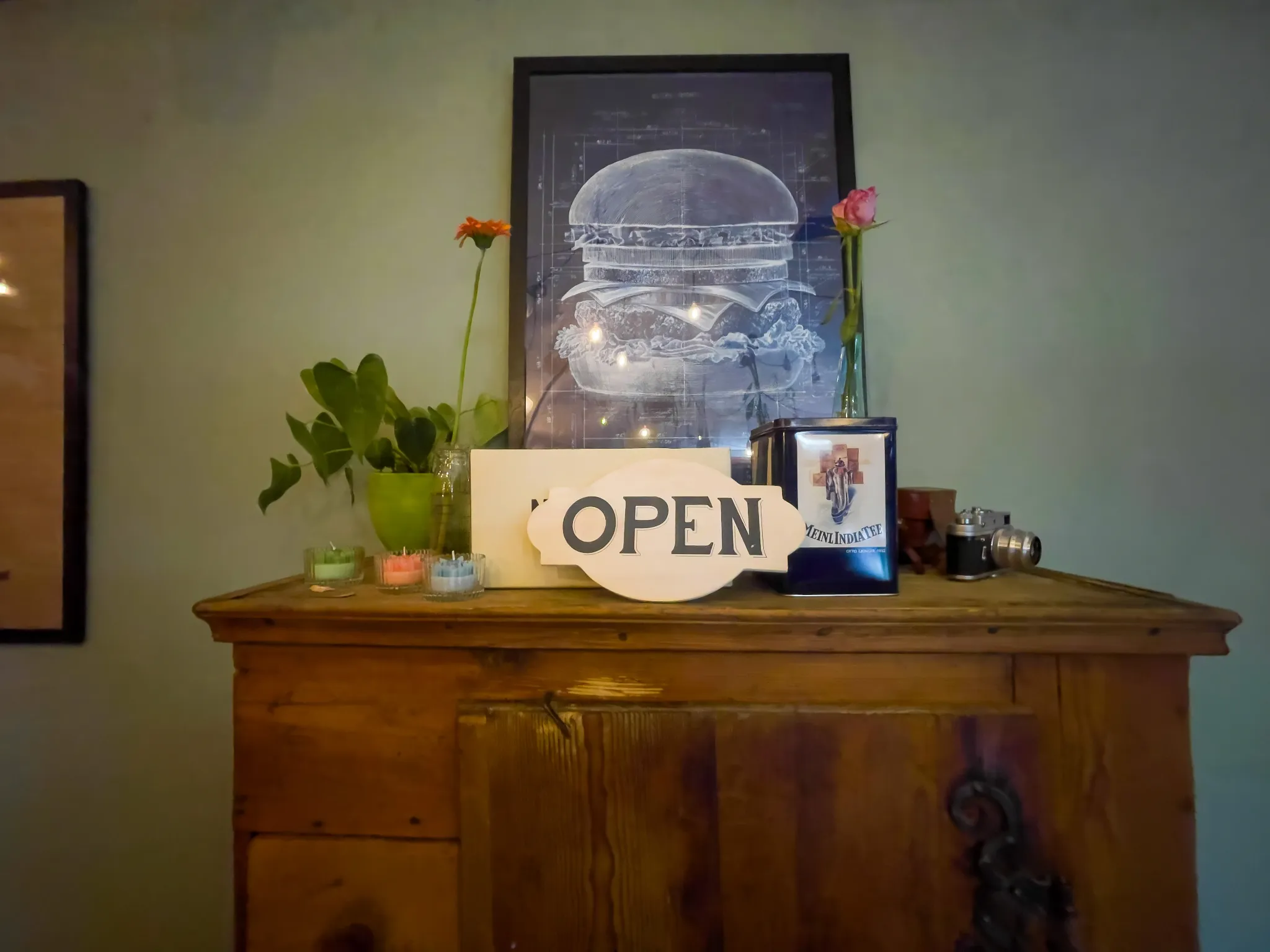 Holzkommode mit Open-Schild, Pflanzen und gerahmtem Motiv